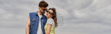 Homme barbu à la mode en lunettes de soleil et gilet en denim regardant petite amie brune dans une robe de soleil élégante et debout avec un ciel nuageux flou en arrière-plan, concept de retraite à la campagne, bannière 