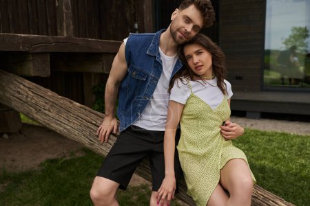Photo for Smiling and stylish bearded man in denim vest hugging brunette girlfriend in sundress and looking at camera while sitting on wooden log in rural setting, countryside retreat concept - Royalty Free Image