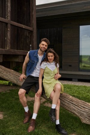 Cheerful and stylish brunette romantic couple in summer outfits and boots hugging while sitting on wooden log in rural setting at background, countryside retreat concept, tranquility