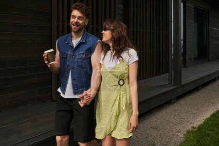Homme barbu souriant et élégant tenant du café à emporter et la main de la petite amie brune en lunettes de soleil et robe d'été tout en marchant près de la maison dans un cadre rural, concept de plaisir en plein air, tranquillité