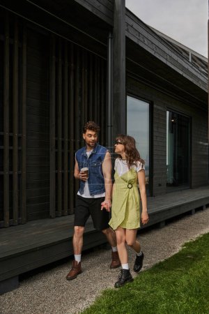 Stylish man in summer outfit holding coffee to go and hand of cheerful girlfriend in sunglasses and sundress while walking near wooden house in rural setting, outdoor enjoyment concept, tranquility