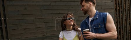 Femme brune souriante en lunettes de soleil et tenue d'été regardant petit ami barbu tenant du café à emporter tout en marchant près de la maison en bois à l'arrière-plan, concept de plaisir en plein air, bannière 