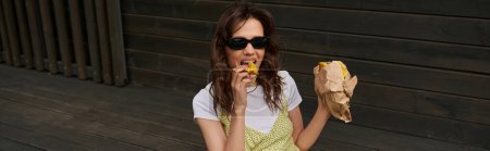 Trendy brunette woman in sunglasses and stylish sundress eating tasty fresh bun while sitting near wooden house in rural setting, summer vibes concept, banner, tranquility