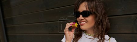 Cheerful brunette woman in sunglasses and summer outfit holding fresh tasty bun while spending time and relaxing near wooden house in rural setting, summer vibes concept, banner 