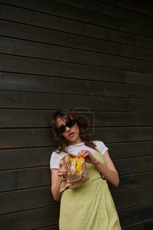 Trendy brunette woman in sunglasses and summer outfit holding fresh bun in craft package while standing near wooden house at background, summer vibes concept, tranquility