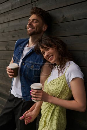 Fröhliche und stylische brünette Frau im Sommeroutfit, die Hand eines bärtigen Freundes in Jeansjacke und Coffee to go hält und in der Nähe eines Holzhauses steht, unbeschwerte Momente.