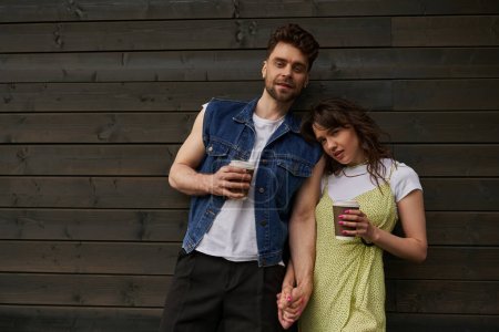 Trendy brunette couple in summer outfits holding hands and takeaway coffee and looking at camera while standing near wooden house in rural setting, carefree moments concept