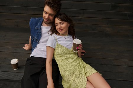 High angle view of relaxed brunette woman in sundress holding coffee to go and sitting near stylish smiling boyfriend and wooden house in rural setting, carefree moments concept, tranquility