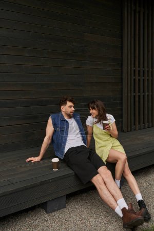 Full length of cheerful brunette woman in stylish summer outfit holding coffee to go and talking to boyfriend while sitting together near wooden house in rural setting, carefree moments concept