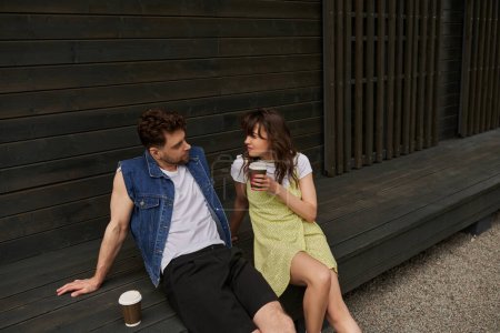 Stilvolle brünette Frau in Sonnenanzüge mit Kaffee zum Mitnehmen und Blick auf bärtigen Freund in Jeansweste, während sie in der Nähe von Holzhaus in ländlicher Umgebung sitzt, unbeschwerte Momente Konzept