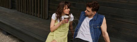 Stylish brunette man in denim vest looking at girlfriend in summer outfit holding coffee to go and sitting near wooden house in rural setting, carefree moments concept, banner 
