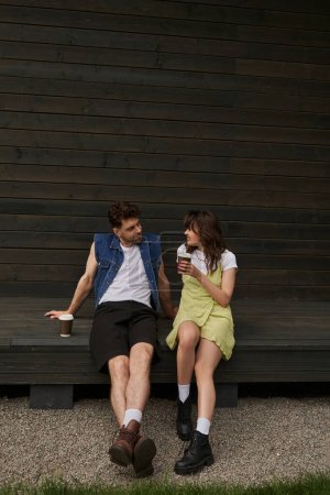Pleine longueur de femme brune joyeuse en bottes et robe de soleil tenant café pour aller et parler au petit ami barbu en gilet de denim tout en étant assis près de la maison en bois, concept d'ambiance sereine