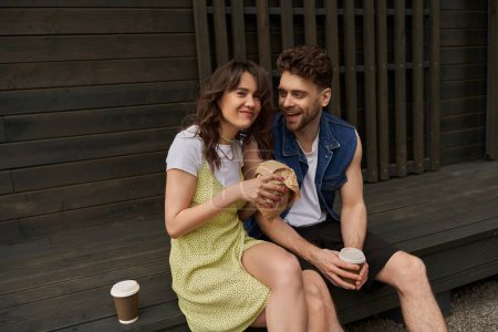 Photo for Cheerful brunette woman in sundress holding craft package and looking at camera near bearded boyfriend with coffee to go and wooden house in rural setting, serene ambiance concept - Royalty Free Image