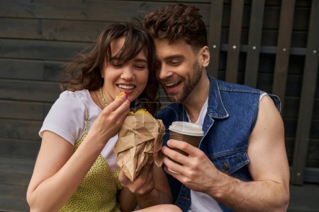 Cheerful romantic couple in summer outfits holding coffee to go and fresh bun in craft package while sitting near wooden house at background, serene ambiance concept, tranquility