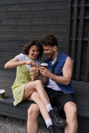 Téléchargez les photos : Femme brune positive en bottes et robe de soleil tenant chignon frais près copain élégant en gilet en denim avec du café à emporter tout en étant assis près de la maison en bois à l'arrière-plan, concept d'ambiance sereine - en image libre de droit
