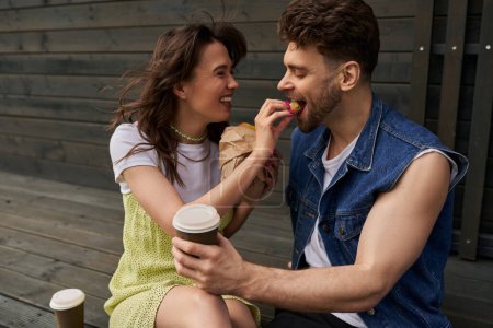 Cheerful and stylish brunette woman in sundress feeding boyfriend with bun and sitting near coffee to go and wooden house at background, serene ambiance concept, tranquility