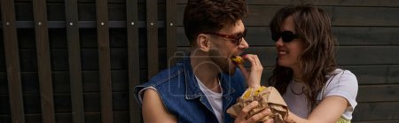 Cheerful brunette woman in sunglasses feeding stylish boyfriend with fresh bun while spending time near wooden house at background in rural setting, serene ambiance concept, banner 