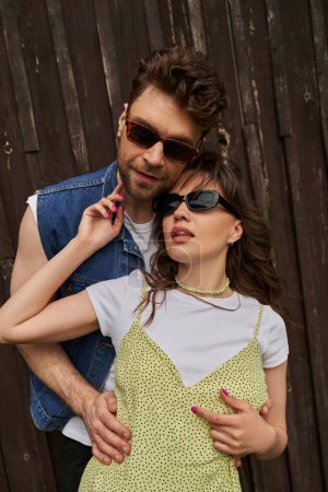 Portrait of stylish bearded man in sunglasses and denim vest embracing brunette girlfriend in sundress and standing together near wooden building outdoors, countryside exploration concept