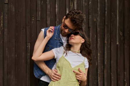 Modisches romantisches Paar in Sonnenbrille und sommerlichem Outfit, das sich umarmt, während es in der Nähe eines Holzhauses steht und Zeit in ländlicher Umgebung verbringt, Konzept zur Erkundung der Landschaft, Ruhe
