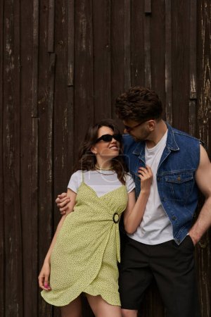 Fashionable bearded man in sunglasses and denim vest hugging cheerful girlfriend in sundress and standing together near wooden house at background, countryside exploration concept