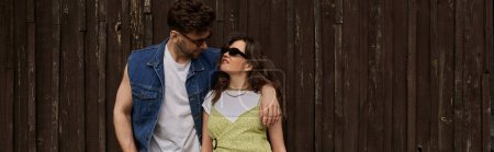 Brunette man in sunglasses and denim vest hugging joyful girlfriend in sundress and standing together near wooden house at background, countryside exploration concept, banner 
