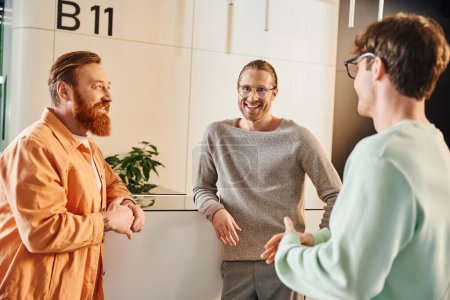optimistic business people in stylish casual clothes talking during meeting in hall of contemporary coworking space, bearded man with colleagues in eyeglasses, startup planning and cooperation