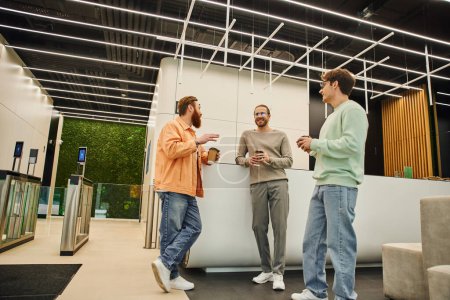 bearded man holding paper cup and talking to smiling colleagues in lobby of contemporary coworking space, meeting of successful businessmen discussing startup project during coffee break