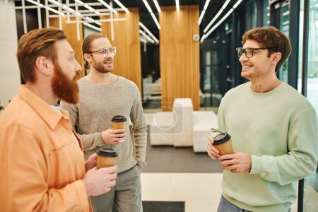 bearded man with stylish and cheerful colleagues in eyeglasses holding takeaway coffee during conversation in lobby of modern coworking space, ambitious businessmen discussing startup project