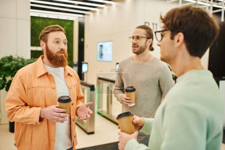 serious bearded man talking to colleagues in eyeglasses holding paper cups with takeaway drinks during coffee break in contemporary coworking office, partnership and collaboration in modern business