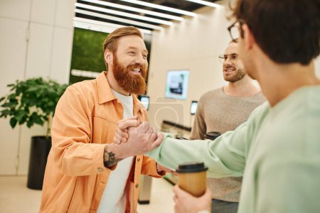 happy bearded entrepreneur shaking hands with business partner near smiling colleague while holding paper cups during coffee break in lobby of modern office space, success and cooperation concept