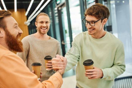 pleased bearded man and his partner in eyeglasses holding takeaway drinks and shaking hands near colleague in hall of modern office, successful entrepreneurs closing deal during coffee break   