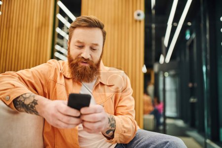 bearded and tattooed businessman in stylish casual clothes networking on mobile phone while sitting on comfortable couch in lounge of modern coworking office on blurred background