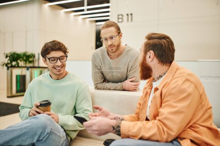 hombre barbudo con teléfono móvil hablando con colegas sonrientes en gafas mientras está sentado en un cómodo sofá durante el descanso de café en el salón de la oficina moderna, empresarios positivos discutiendo startup