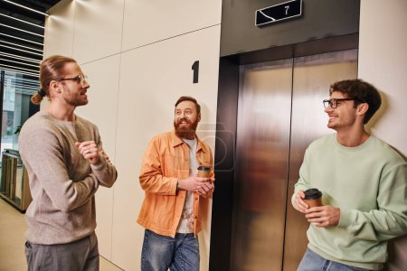 zufriedene Geschäftskollegen in stylischer Freizeitkleidung mit Imbissgetränken in Pappbechern beim Warten auf den Fahrstuhl, erfolgreiche Unternehmer, die sich in der Kaffeepause im modernen Büro unterhalten