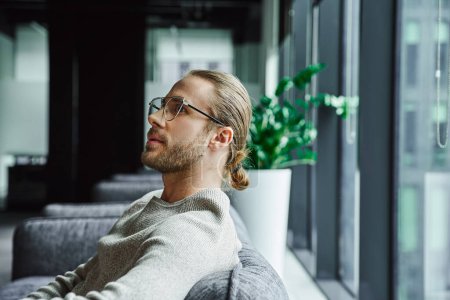Photo for Side view of pensive businessman in stylish eyeglasses and casual clothes thinking on startup project while sitting in office lounge in contemporary coworking space, ambition and creative thinking - Royalty Free Image