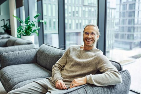 Photo for Happy entrepreneur in stylish eyeglasses and casual clothes, with radiant smile, sitting on comfortable couch near large windows in office lounge and looking at camera, business success concept - Royalty Free Image