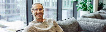 professional headshot of smiling and successful businessman in casual clothes and stylish eyeglasses sitting on comfortable couch in lounge of modern coworking office and looking at camera, banner