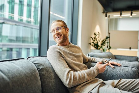 joyful entrepreneur in eyeglasses and casual clothes holding smartphone and looking away through window while sitting on comfortable sofa in modern coworking environment, successful business concept