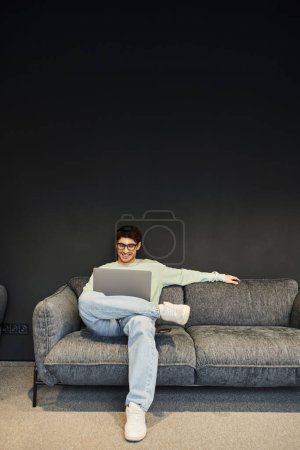 smiling and successful businessman in eyeglasses and stylish casual clothes looking at laptop while sitting on grey couch near black wall in lounge of coworking office with modern interior