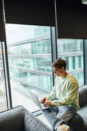 joyful businessman in casual clothes and eyeglasses typing on laptop while sitting on comfortable couch near large windows in lounge of coworking office, modern workplace, business productivity