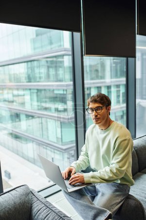 ambitious and career oriented businessman in stylish eyeglasses and casual clothes sitting on comfortable couch with laptop and looking at camera near large windows in lounge of modern office
