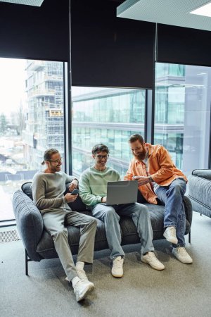 full length of business partners with takeaway drink and mobile phone smiling near colleague working on laptop, sitting on comfortable sofa in lounge of modern coworking office with large windows 