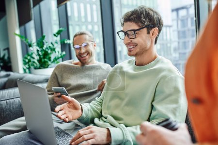 happy entrepreneur in eyeglasses pointing at laptop while working on startup project near business partner with smartphone smiling on blurred background in modern coworking space