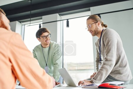 positive architectural designers in casual clothes looking at each other near laptop and businessman on blurred foreground while working on startup project in contemporary coworking office