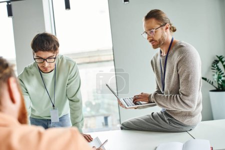 Photo for Architectural designer in eyeglasses and casual clothes sitting on work desk, typing on laptop and looking at colleagues working with drafting in contemporary coworking office, blurred foreground - Royalty Free Image