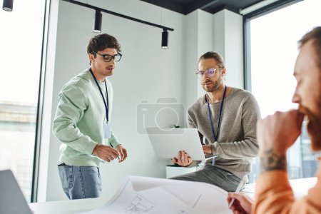 Photo for Thoughtful architectural designer in eyeglasses working on laptop near thoughtful business colleagues and blueprint on blurred foreground, creative coworking in modern office environment - Royalty Free Image