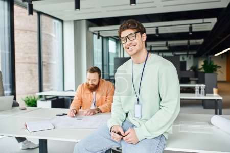 joyful entrepreneur in eyeglasses and casual clothes sitting on work desk and looking at camera while bearded architect working with blueprint in modern coworking space of design studio