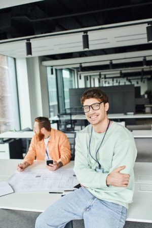 satisfied entrepreneur in eyeglasses sitting on work desk with folded arms and smiling at camera next to bearded architect holding smartphone and working with blueprint on blurred background