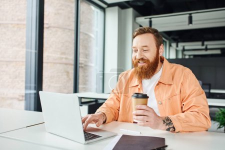 cheerful, tattooed and bearded entrepreneur holding paper cup with coffee to go and networking on laptop near folder on work desk in contemporary coworking office, successful business concept