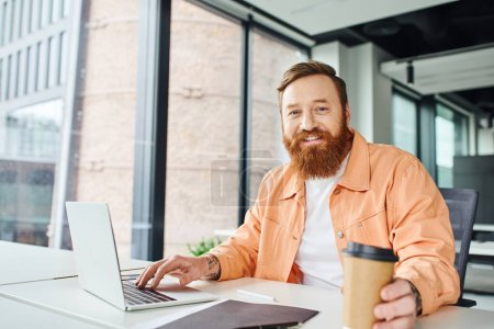 positive emotion of bearded and tattooed man with takeaway drink sitting at workplace near folder and laptop and smiling at camera in contemporary office environment, business success concept